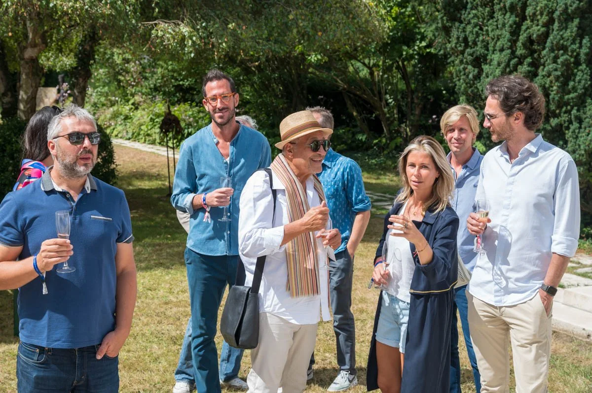 Groupe de personnes buvant du champagne lors d'un rassemblement en plein air sur une pelouse, entouré d'arbres et de verdure. Certains portent des vêtements décontractés, d'autres portent des lunettes de soleil et un chapeau. L'ambiance est festive e