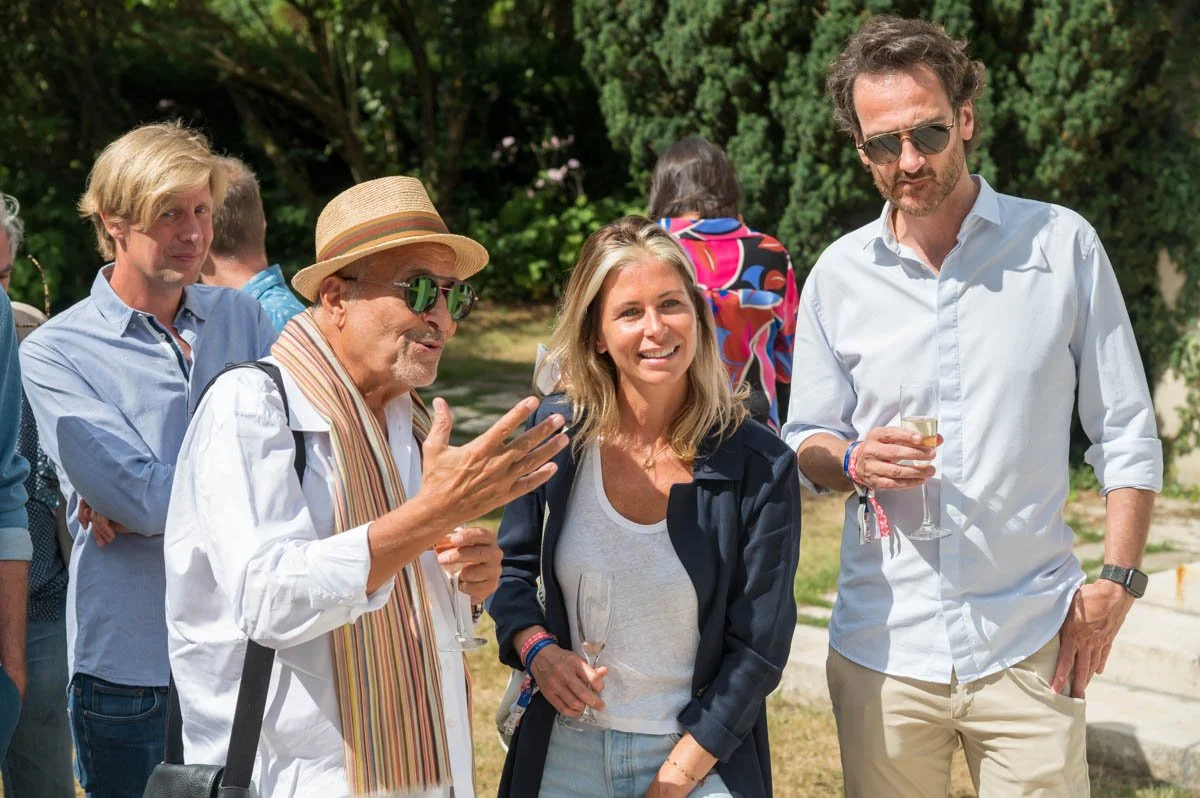Un groupe de personnes lors d'un événement en plein air, certaines tenant des verres. Un homme avec un chapeau et des lunettes parle, une femme et un homme avec des lunettes de soleil écoutent."