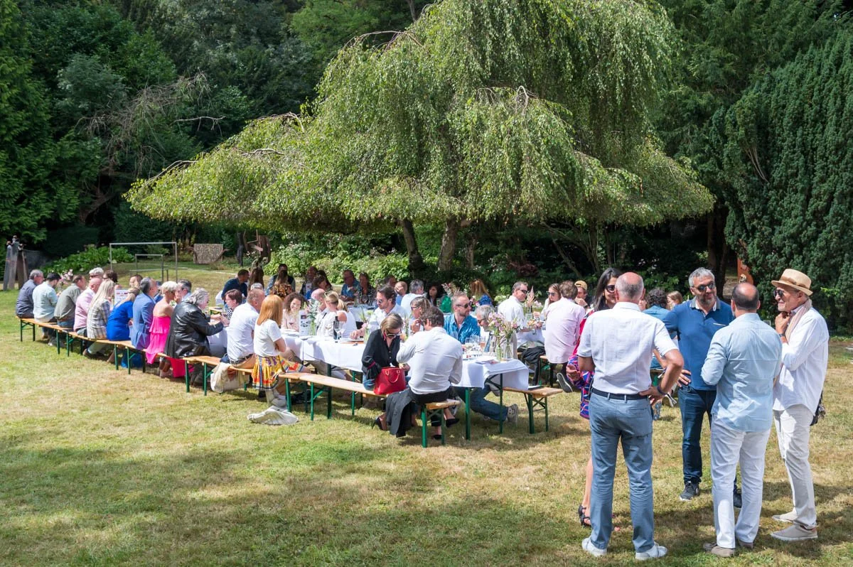 Un grand groupe de personnes assises à des tables de pique-nique en plein air dans un jardin, sous un arbre, pendant un repas. Des gens aussi debout discutent en arrière-plan. Il fait beau et l'endroit est entouré de verdure.