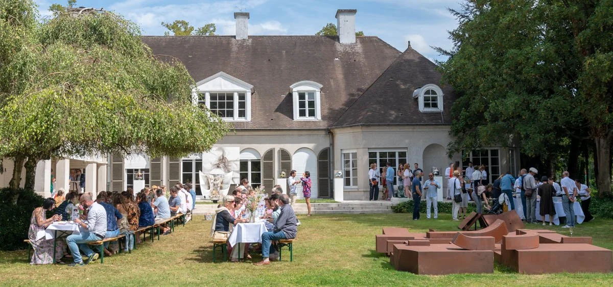 Personnes assises à des tables dans un jardin devant une grande maison de campagne, lors d'un événement social par beau temps.