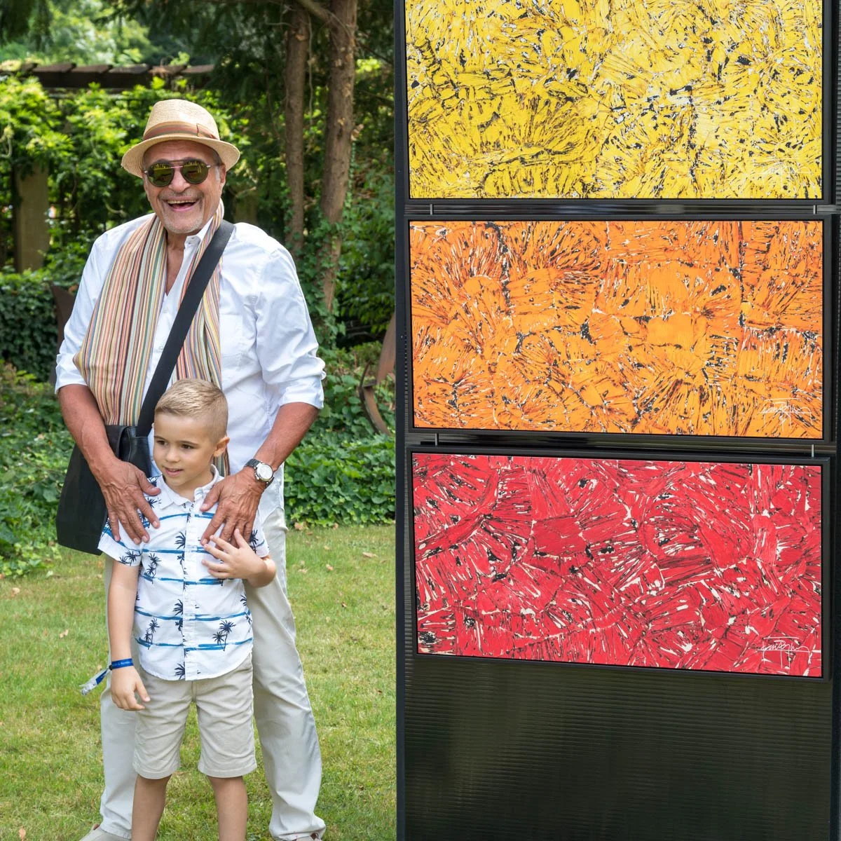 Un homme âgé souriant porte un chapeau et des lunettes de soleil, debout avec un jeune garçon devant un jardin verdoyant. À côté, il y a un panneau avec trois peintures abstraites colorées en jaune, orange et rouge. L'homme porte une écharpe et a un