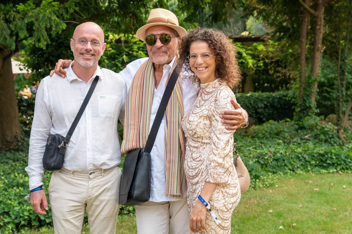 Trois personnes souriantes posent ensemble à l'extérieur, sous des arbres. Elles portent des vêtements estivaux et des sacs en bandoulière.