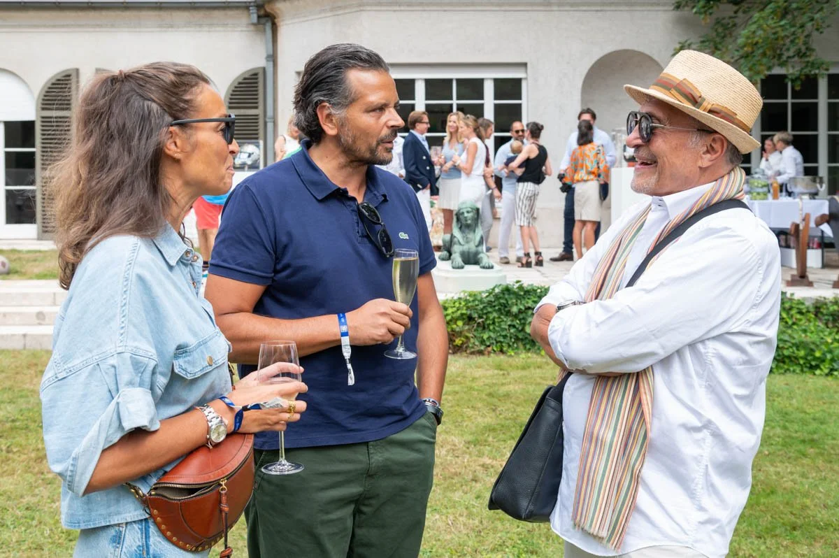 Trois personnes discutant à l'extérieur lors d'un événement social, avec des boissons à la main, entourées d'autres participants à l'arrière-plan.