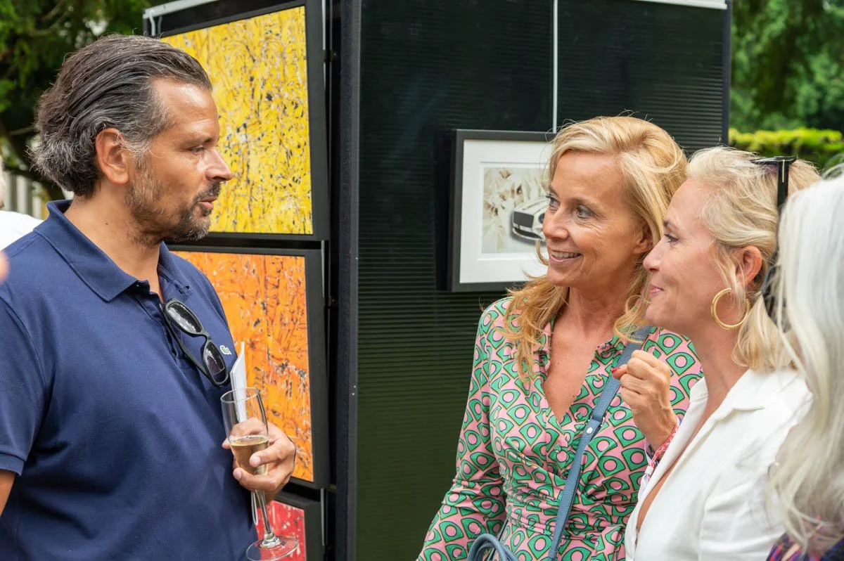 Trois personnes discutent devant des œuvres d'art lors d'une exposition en plein air. Un homme tient un verre de champagne tandis que deux femmes lui parlent en souriant. Les œuvres d'art montrent des motifs colorés.