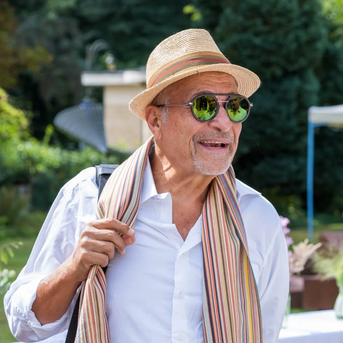 Un homme portant un chapeau de paille et des lunettes de soleil réfléchissantes, vêtu d'une chemise blanche et écharpe à rayures, sourit à l'extérieur avec un fond flou de verdure.