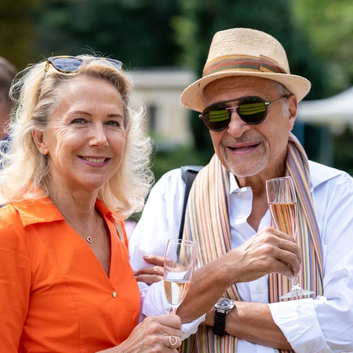 Un homme et une femme âgés souriant, tenant des verres de champagne. L'homme porte un chapeau et des lunettes de soleil, la femme porte une chemise orange. Ils se trouvent à l'extérieur.