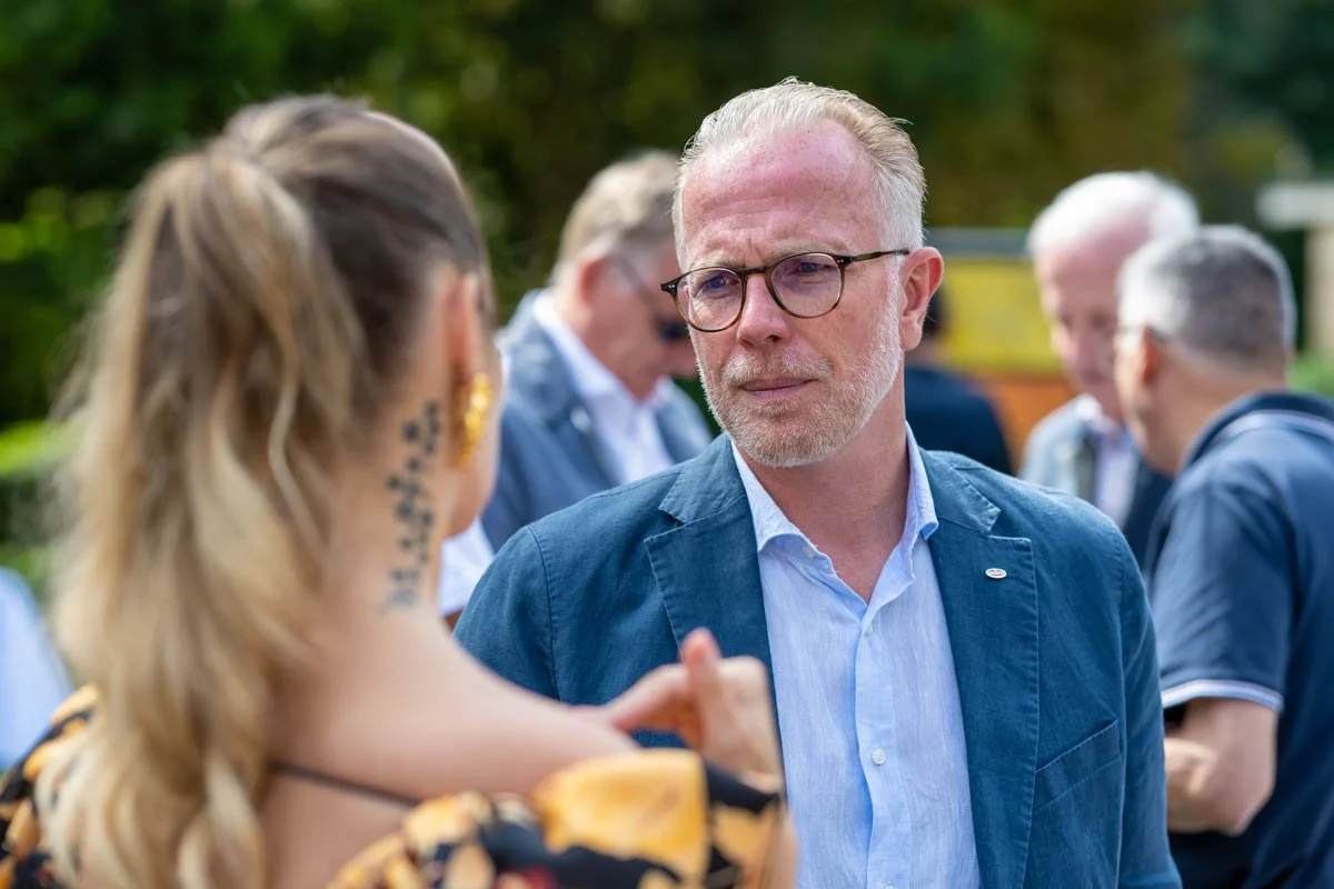 Une personne avec des lunettes parle à une femme blonde avec un tatouage sur le cou, en arrière-plan plusieurs personnes floues.