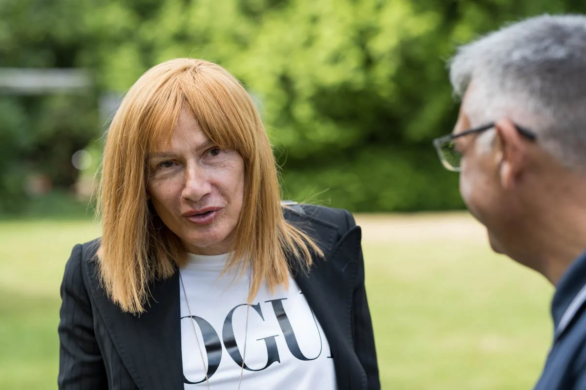 Deux personnes discutant à l'extérieur, l'une portant une veste noire et un t-shirt avec le mot "Vogue".