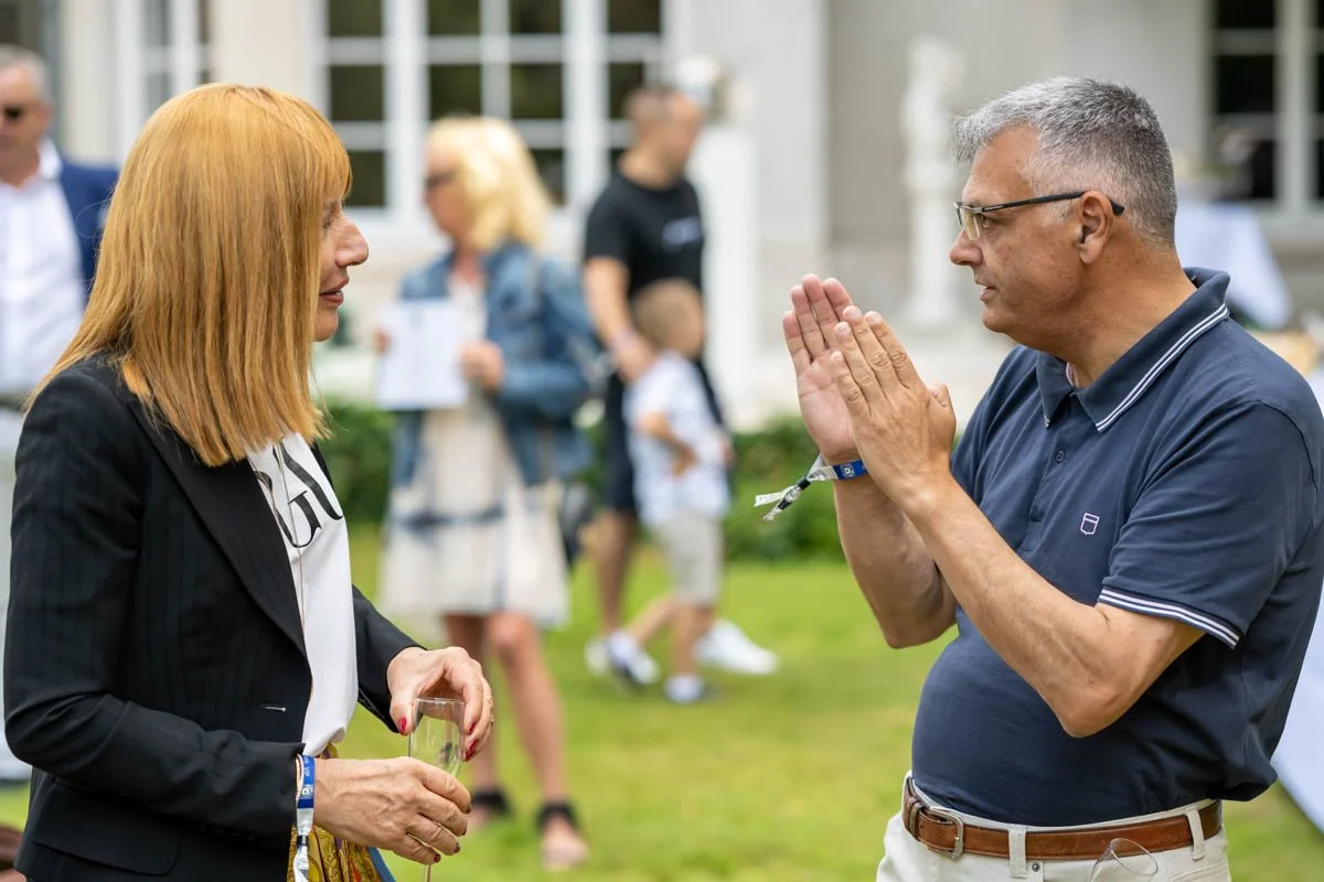Deux personnes discutant à l'extérieur lors d'une rencontre sociale, l'une tenant un verre et l'autre gesticulant. Des personnes floues et un jardin en arrière-plan.