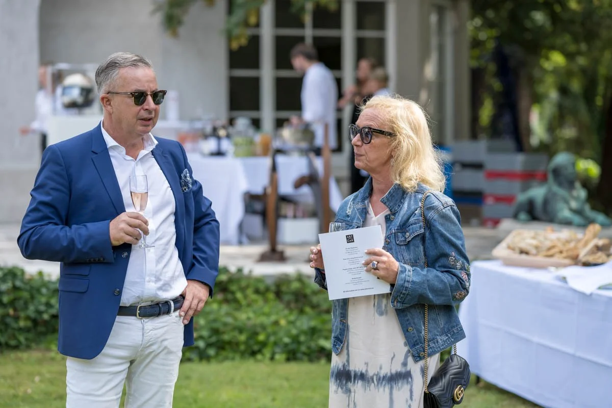 Deux personnes discutent lors d'un événement en plein air, un homme tenant un verre de champagne et une femme portant des lunettes de soleil et un dépliant. Il y a des tables avec de la nourriture à l'arrière-plan.