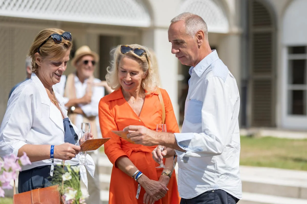 Trois personnes discutant en extérieur lors d'un événement, tenant des cartes et des verres de champagne. La femme au milieu porte une robe orange.