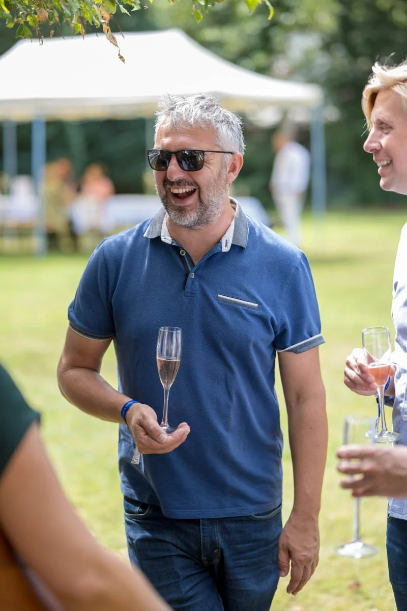 Un homme souriant portant des lunettes de soleil et un polo bleu tient un verre de champagne dans un jardin, discutant avec d'autres personnes pendant une fête en plein air.