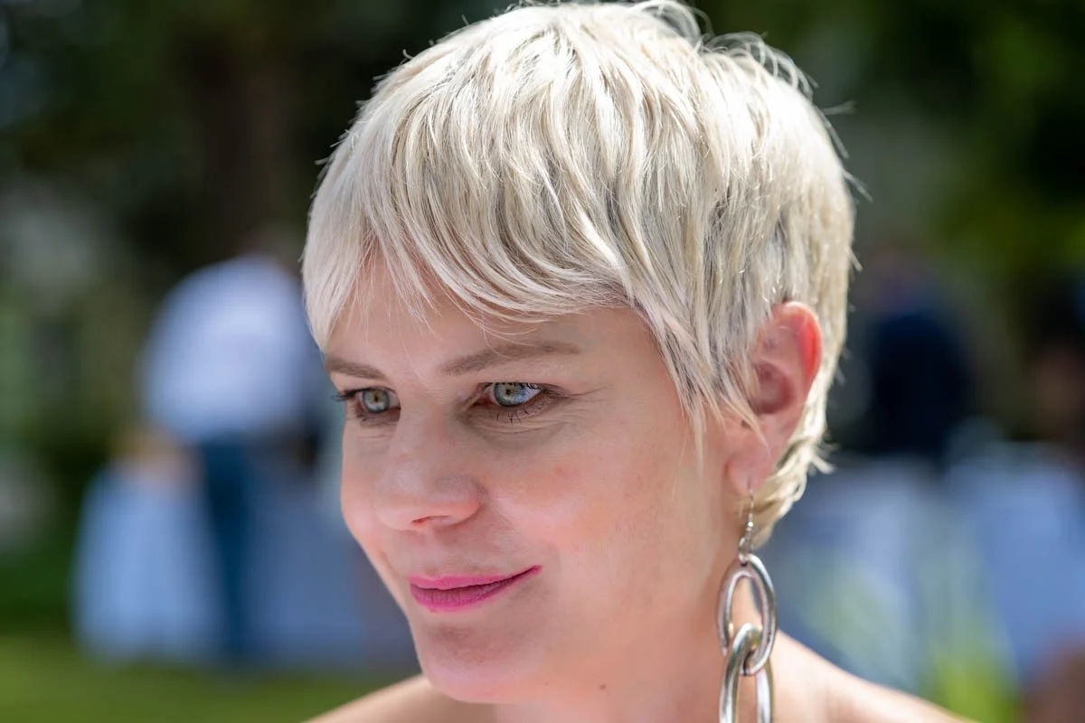 Femme souriante aux cheveux courts et blonds, portant des boucles d'oreilles en métal, avec un fond flou extérieur.