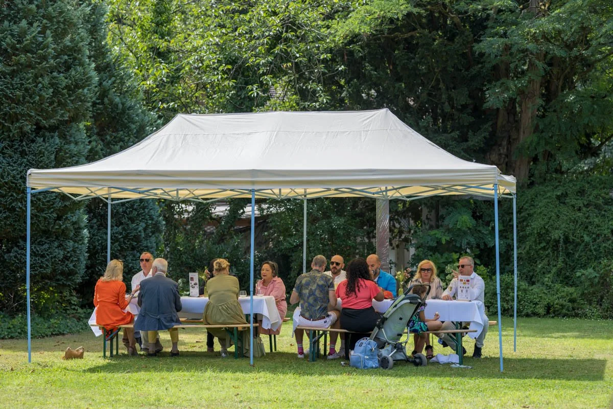 Groupe de personnes assis sous une tente blanche dans un parc, discutant et partageant un repas sur une grande table rectangulaire, avec des arbres en arrière-plan.
