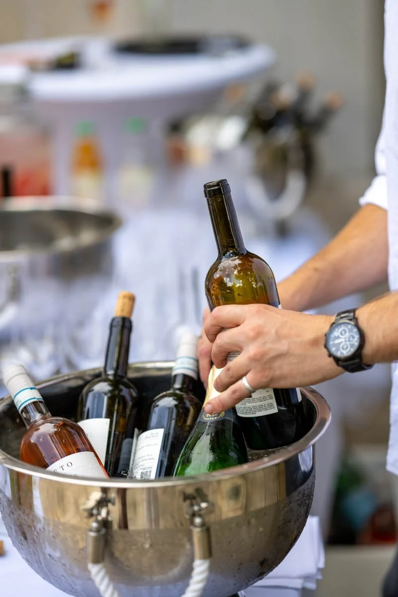 Une personne tient une bouteille de vin à moitié sortie d'un seau en métal contenant plusieurs autres bouteilles de vin, sur une table lors d'une fête ou événement.