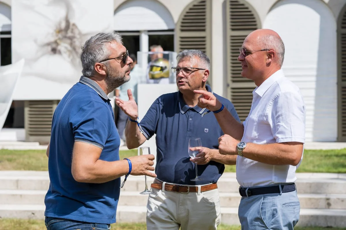 Trois hommes discutant à l'extérieur avec des verres à la main, devant un bâtiment aux volets fermés.
