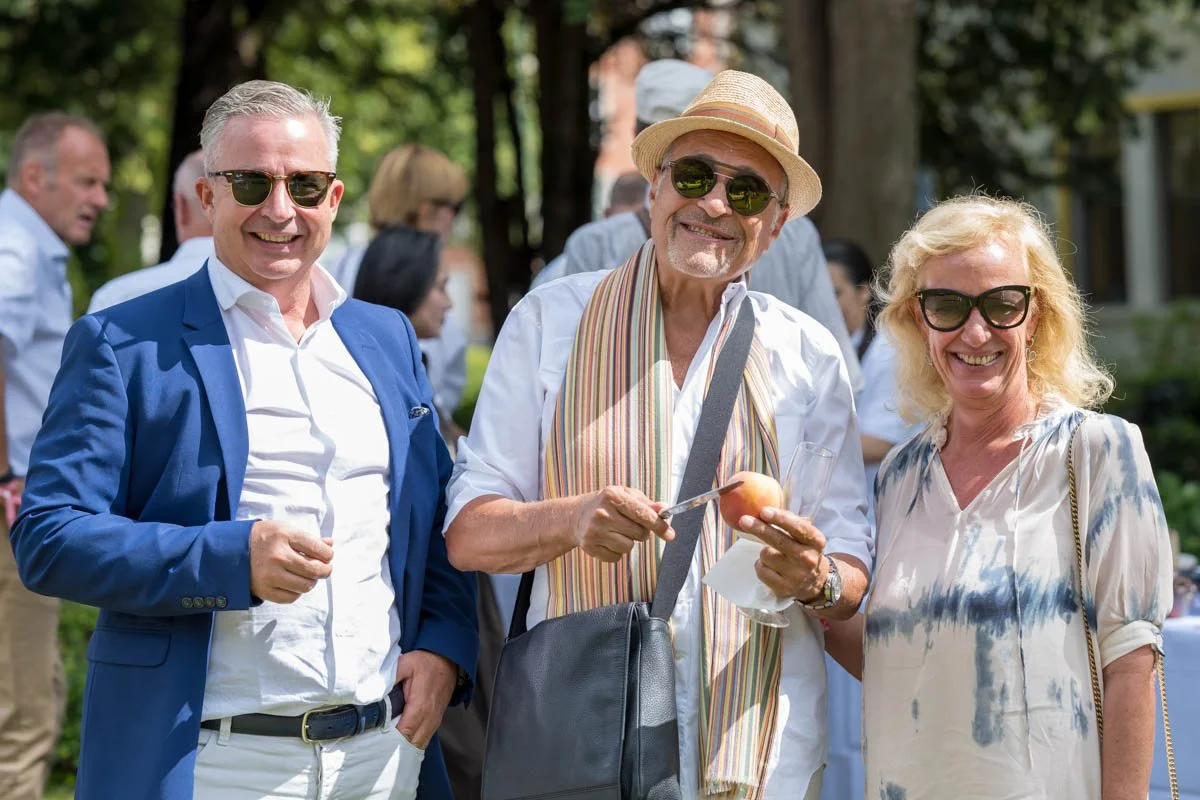 Trois personnes souriantes portant des lunettes de soleil à l'extérieur, un homme au centre avec un chapeau et une écharpe, tenant une pêche et un couteau. Il y a des gens en arrière-plan.