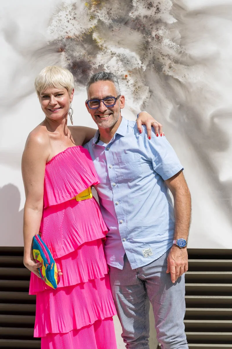 Un homme et une femme posant ensemble, la femme portant une robe rose et tenant un sac à main coloré, l'homme portant une chemise bleue et des lunettes.