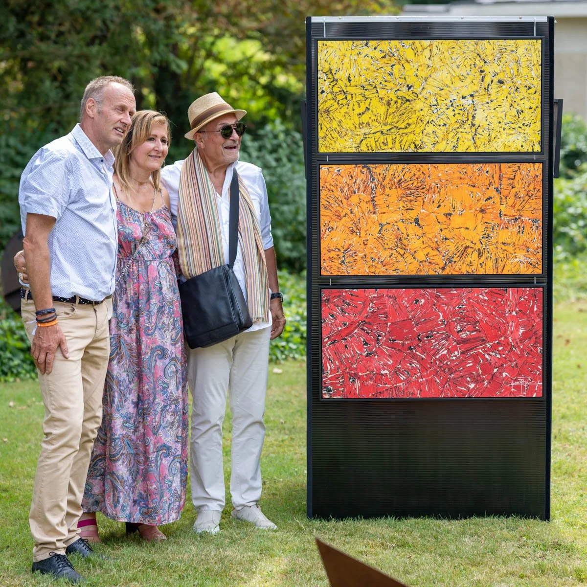 Trois personnes posent devant une œuvre d'art colorée composée de trois panneaux: jaune, orange, et rouge, dans un jardin verdoyant.
