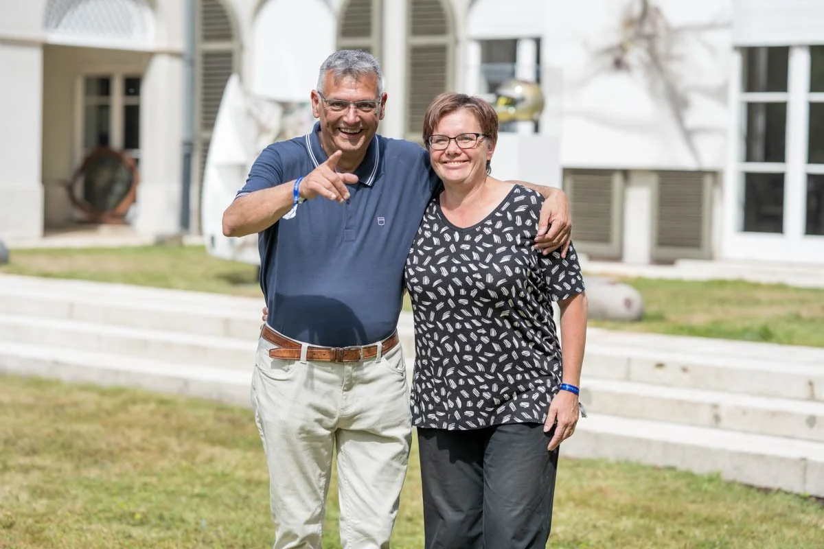 Un homme et une femme souriants s'embrassent à l'extérieur, dans un jardin, devant un bâtiment. L'homme porte un polo bleu et un pantalon clair, tandis que la femme porte un t-shirt à motifs noirs et blancs et un pantalon foncé.