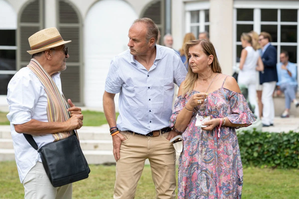 Trois personnes discutant en plein air lors d'un événement, avec d'autres participants flous à l'arrière-plan.