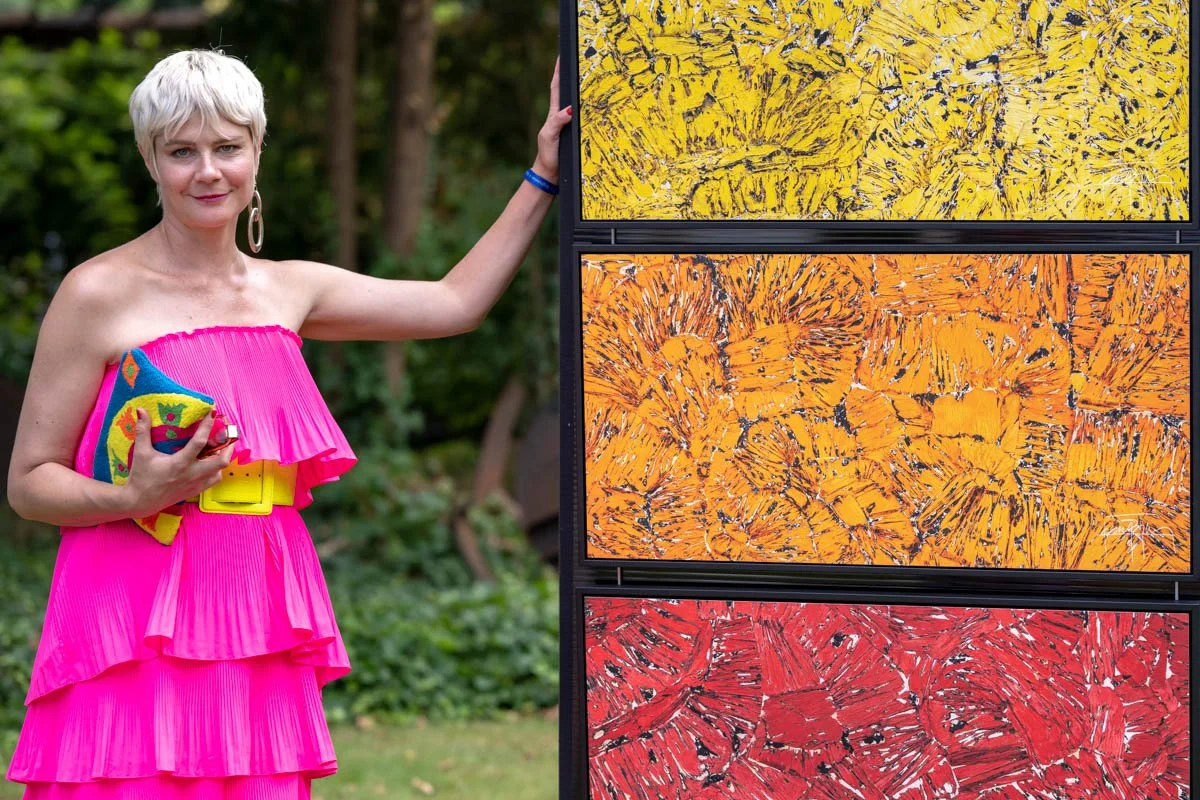Une femme en robe rose vif pose à côté de trois œuvres d'art abstrait en jaune, orange et rouge. Elle tient une pochette colorée et se trouve dans un jardin.