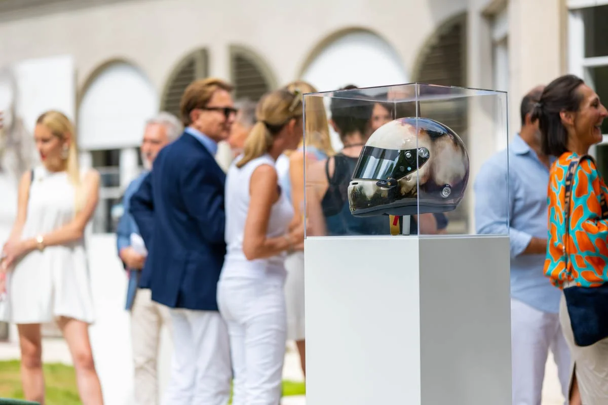 Casque de moto exposé dans une boîte en verre, avec des personnes en arrière-plan lors d'un événement en plein air.