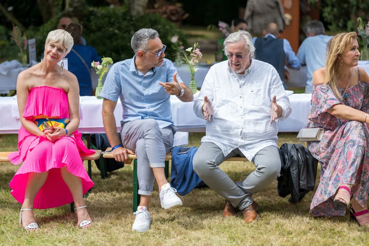 Groupe de personnes assises sur un banc lors d'un événement en plein air, avec des fleurs et des tables en arrière-plan.