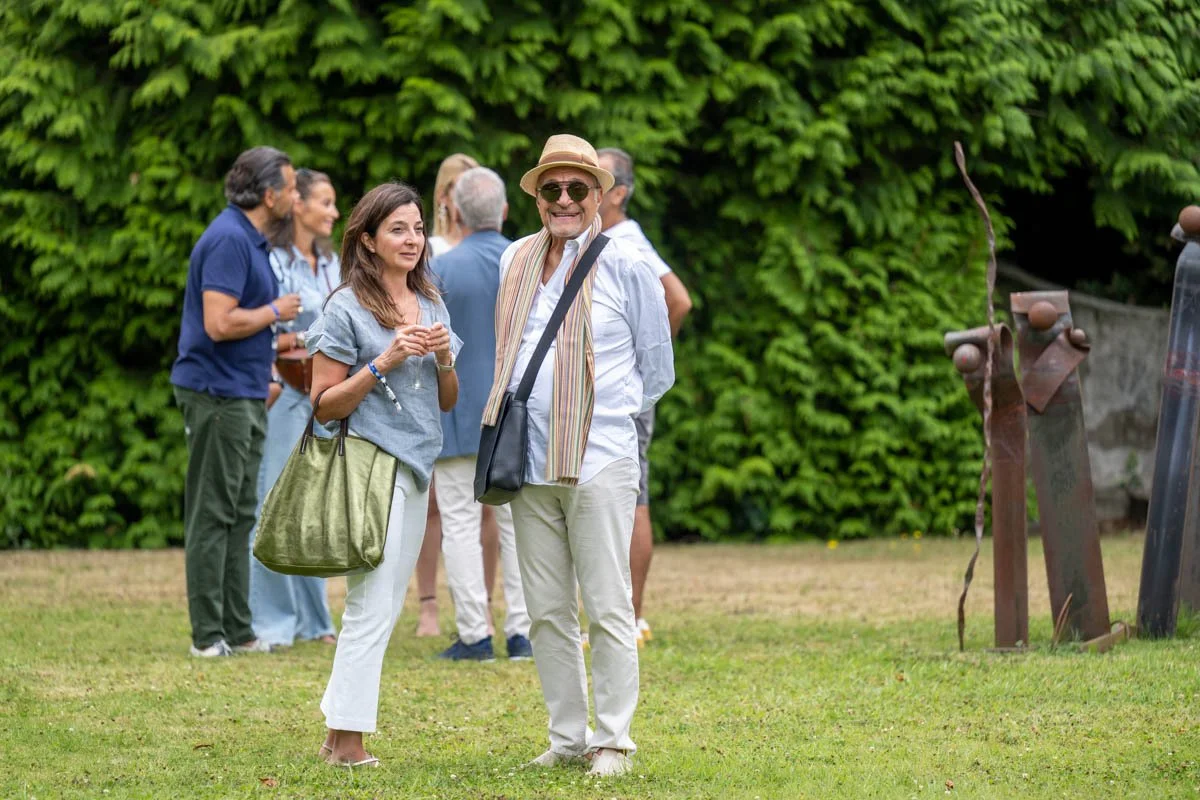 Un groupe de personnes se tient sur une pelouse, discutant et socialisant, avec des statues métalliques à côté. En arrière-plan, il y a une haie de buissons verts.
