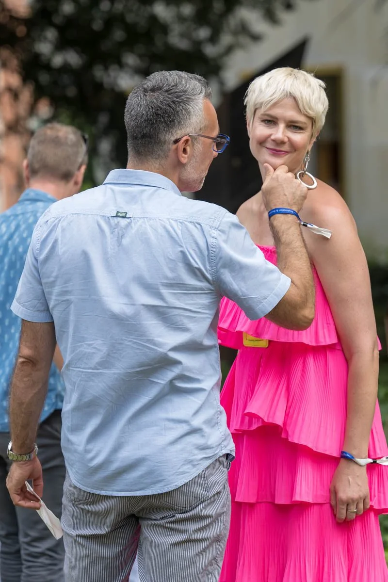 Un homme en chemise bleue ajuste un vêtement d'une femme en robe rose vif lors d'un événement en extérieur.