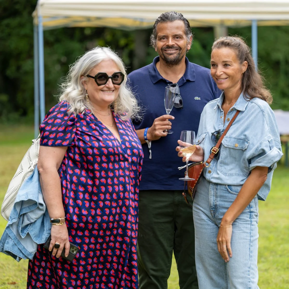 Trois personnes posant lors d'un événement en plein air. L'une d'elles porte une robe bleue à motifs, une autre un polo marine et lunettes de soleil, et la troisième une tenue en jean. Ils tiennent des verres, suggérant une ambiance festive ou décont