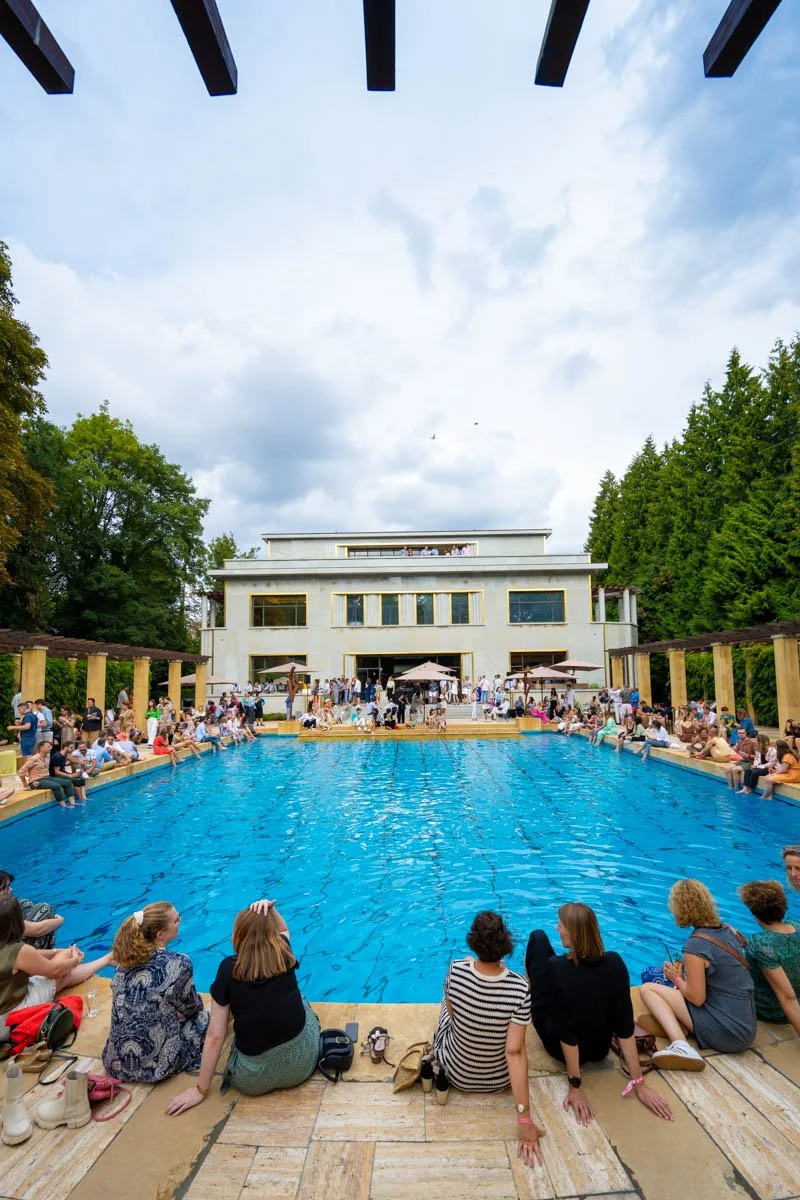 Des personnes assises autour d'une grande piscine extérieure, avec un bâtiment moderne en arrière-plan et entouré de verdure.