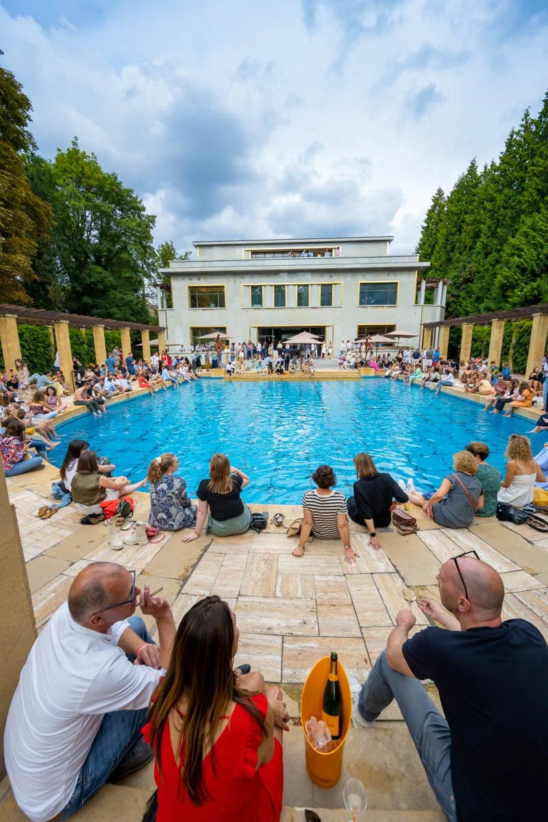 Un groupe de personnes assises autour d'une grande piscine en extérieur, devant un bâtiment moderne avec plusieurs étages. Il y a des arbres et un ciel partiellement nuageux en arrière-plan. Certaines personnes tiennent des verres, indiquant un événe