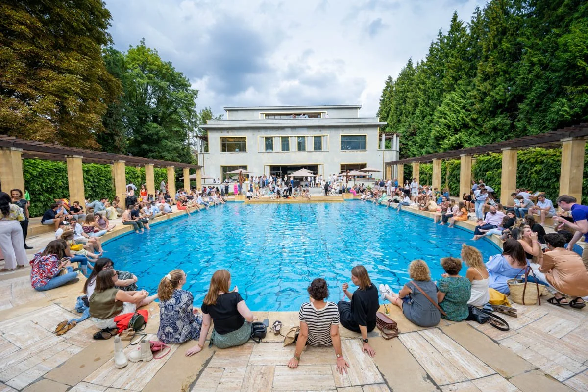 Des gens assis autour d'une piscine bleue, devant un bâtiment moderne, entouré d'arbres.