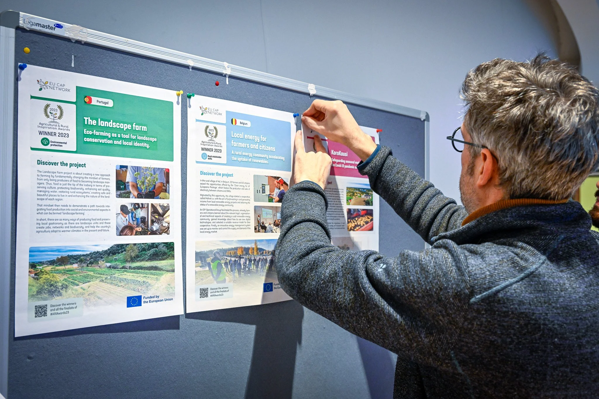 Un homme accrochant des affiches sur un tableau d'affichage, présentant des projets liés à l'agriculture et l'énergie durable, avec des mentions de réseaux européens et financements de l'Union Européenne.