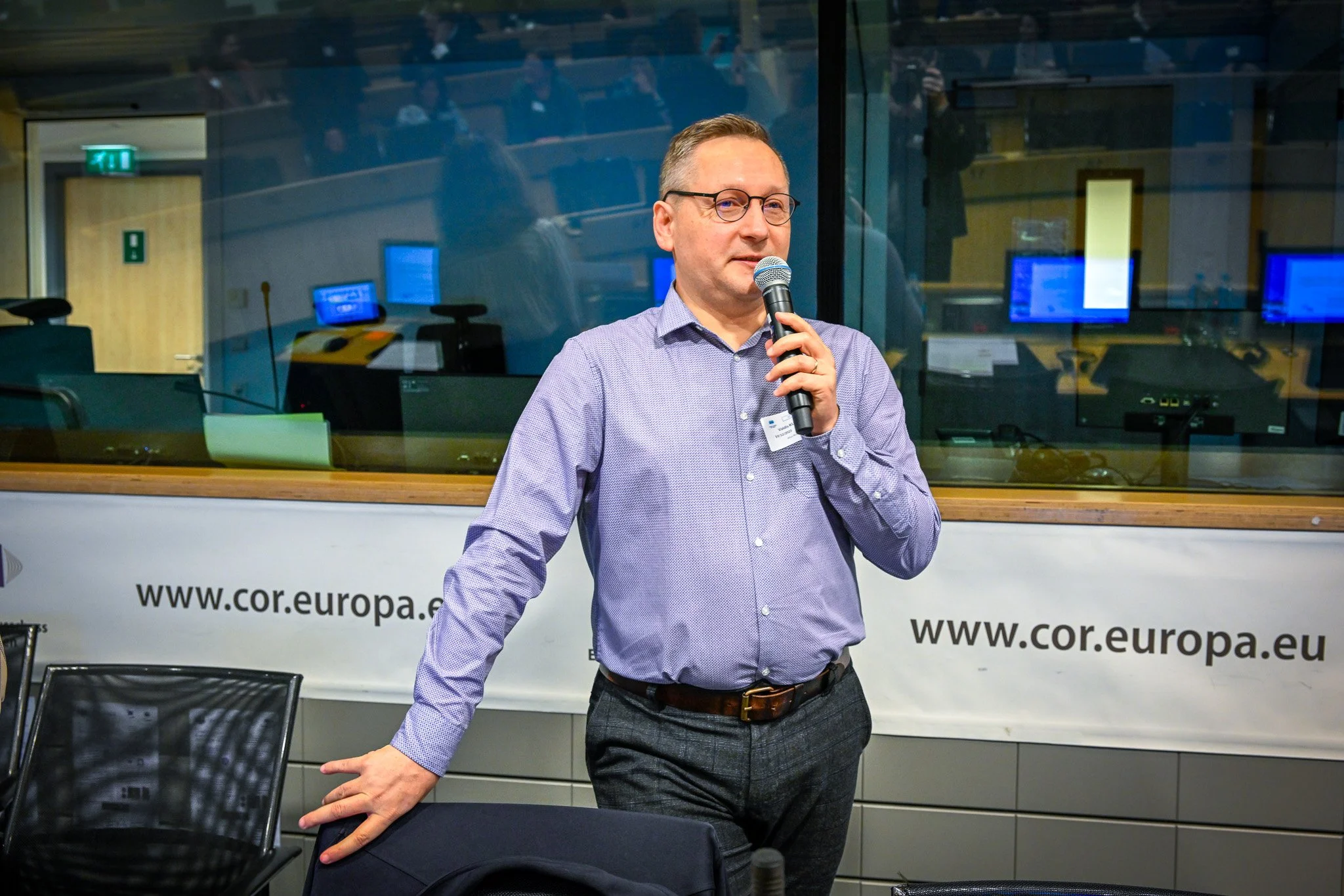 Un homme en chemise bleue parle dans un microphone lors d'une conférence, debout devant un panneau indiquant "cor.europa.eu".