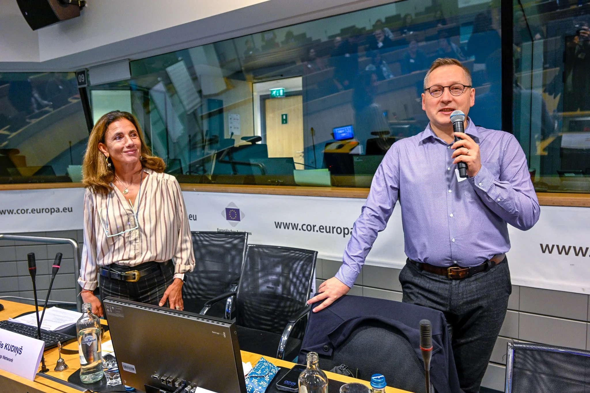 Un homme tenant un microphone et une femme debout dans une salle de conférence moderne avec des ordinateurs et des micros, et une bannière du site web cor.europa.eu en arrière-plan.