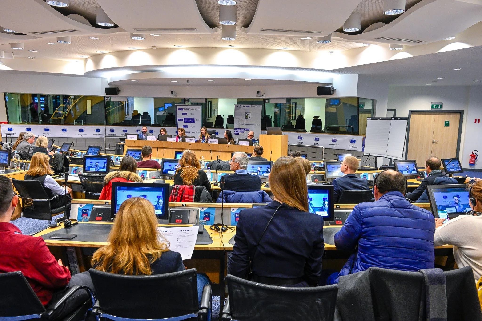 Salle de conférence avec des participants assis et des écrans, réunion en cours.
