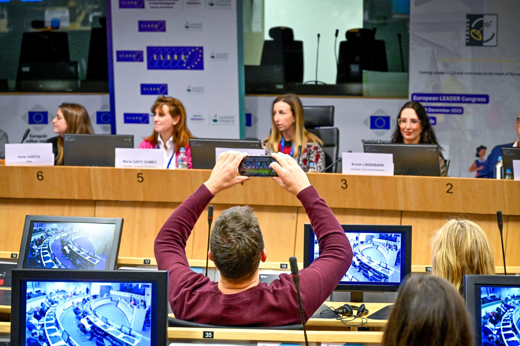 Une conférence avec plusieurs personnes assises à une table, des écrans d'ordinateur affichant la salle, et un participant prenant une photo avec un smartphone.