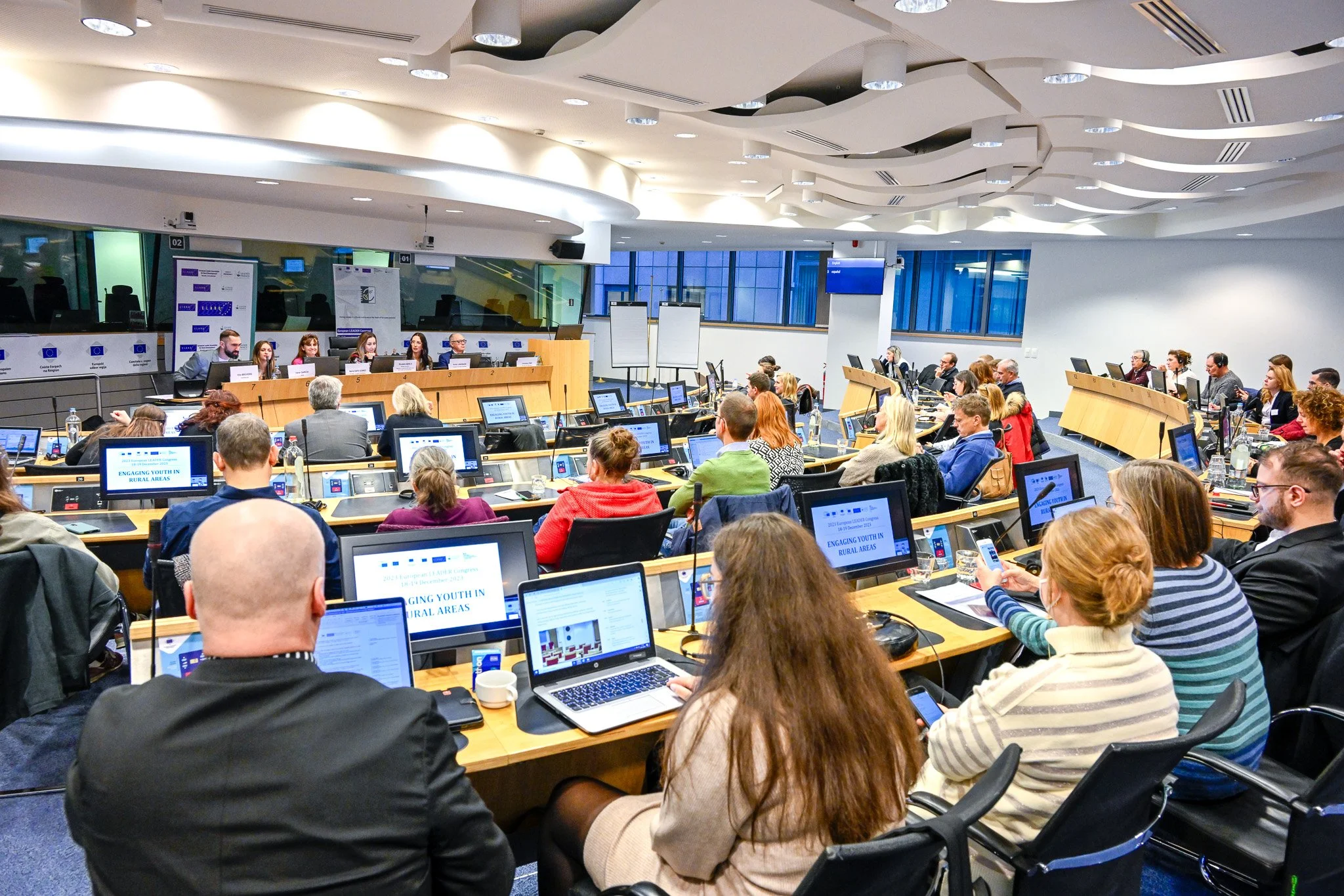Salle de conférence avec des participants assis autour de tables équipées d'ordinateurs, écoutant un panel de conférenciers devant la salle.