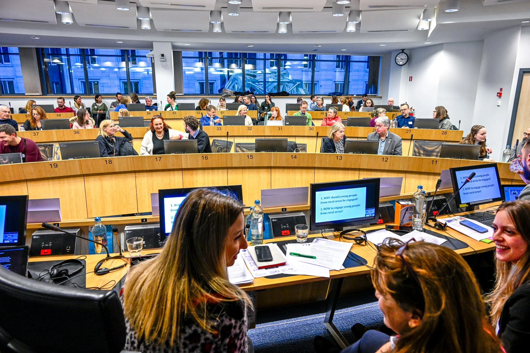 Salle de conférence avec des personnes assises à des bureaux numérotés, discutant et regardant des écrans avec des questions sur l'engagement des jeunes des zones rurales.