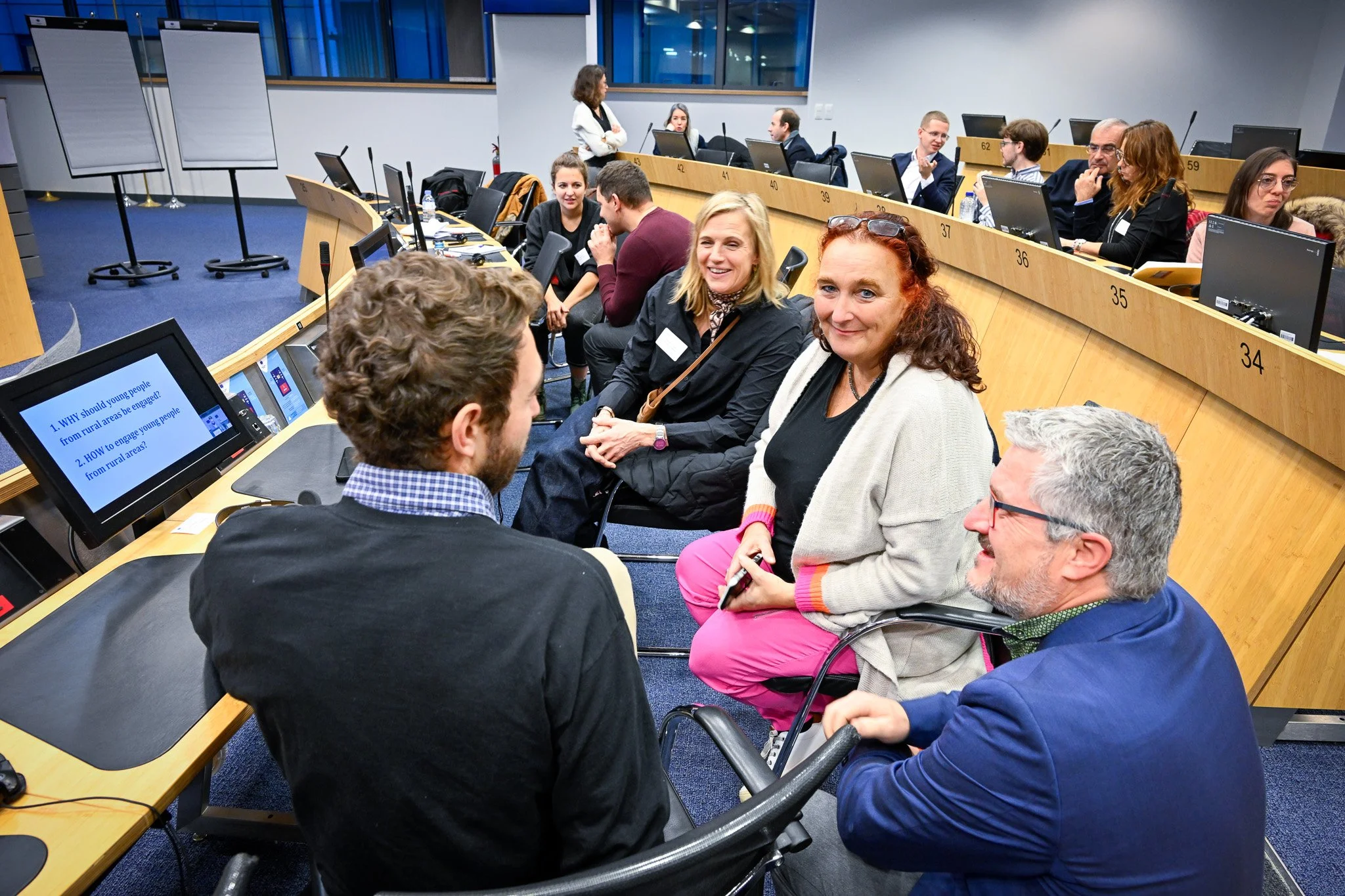 Personnes discutant dans une salle de conférence avec des ordinateurs et écrans, tableau numérique montrant des questions sur les jeunes dans les zones rurales, sièges numérotés et plusieurs participants.