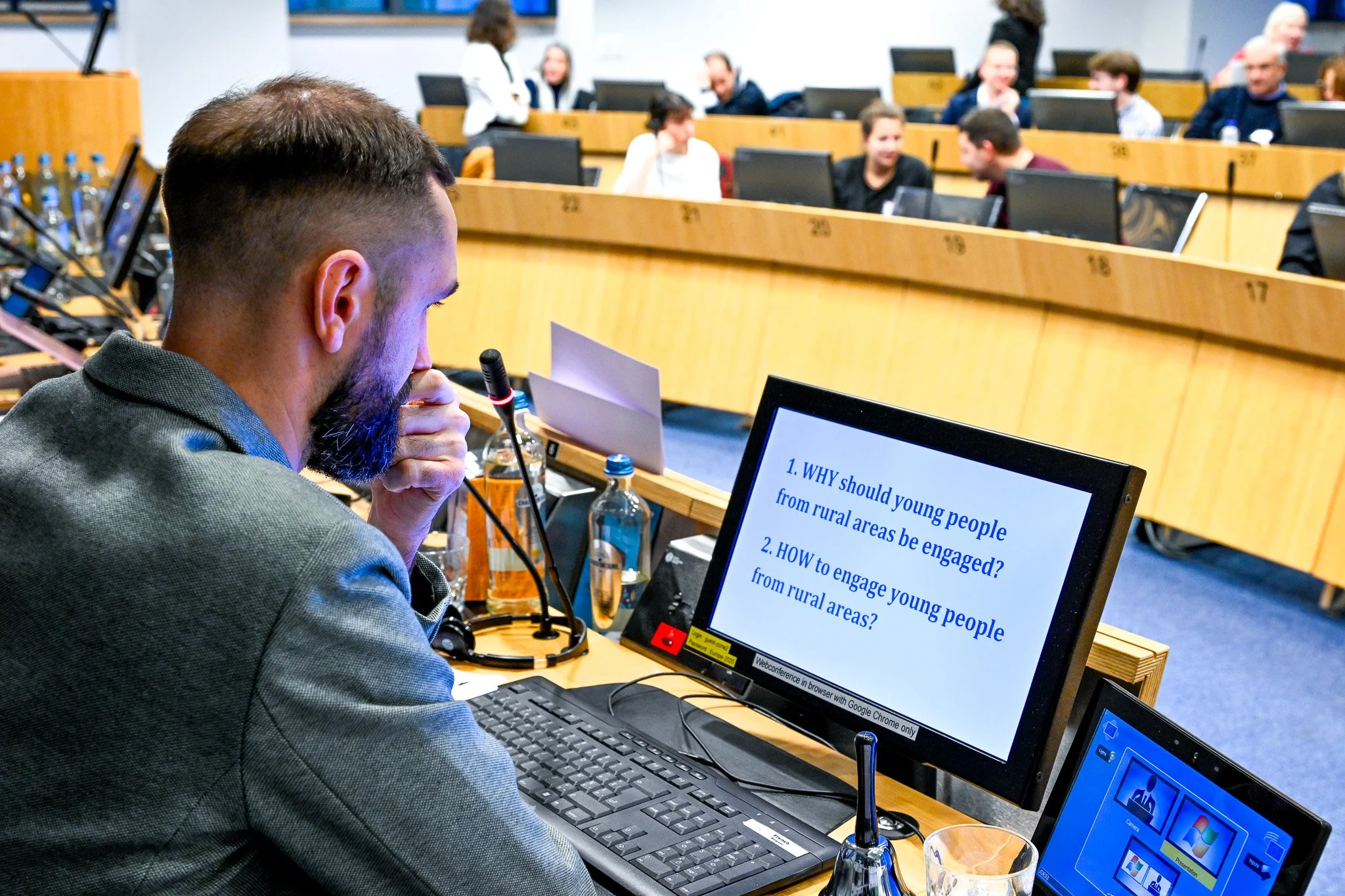 Un homme assis devant un écran d'ordinateur dans une salle de conférence, avec un texte sur l'écran demandant l'engagement des jeunes des zones rurales. D'autres personnes sont assises à l'arrière-plan, participant à une discussion.