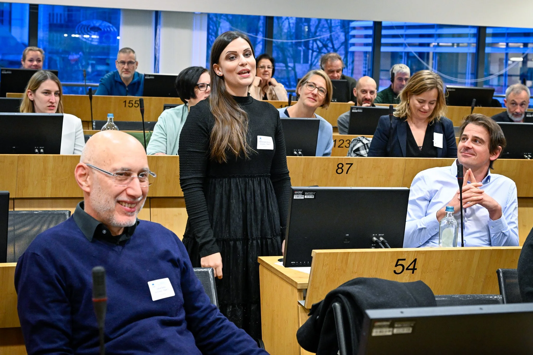 Groupe de personnes dans une salle de conférence, certaines assises et d'autres debout, avec des écrans d'ordinateur sur les bureaux.