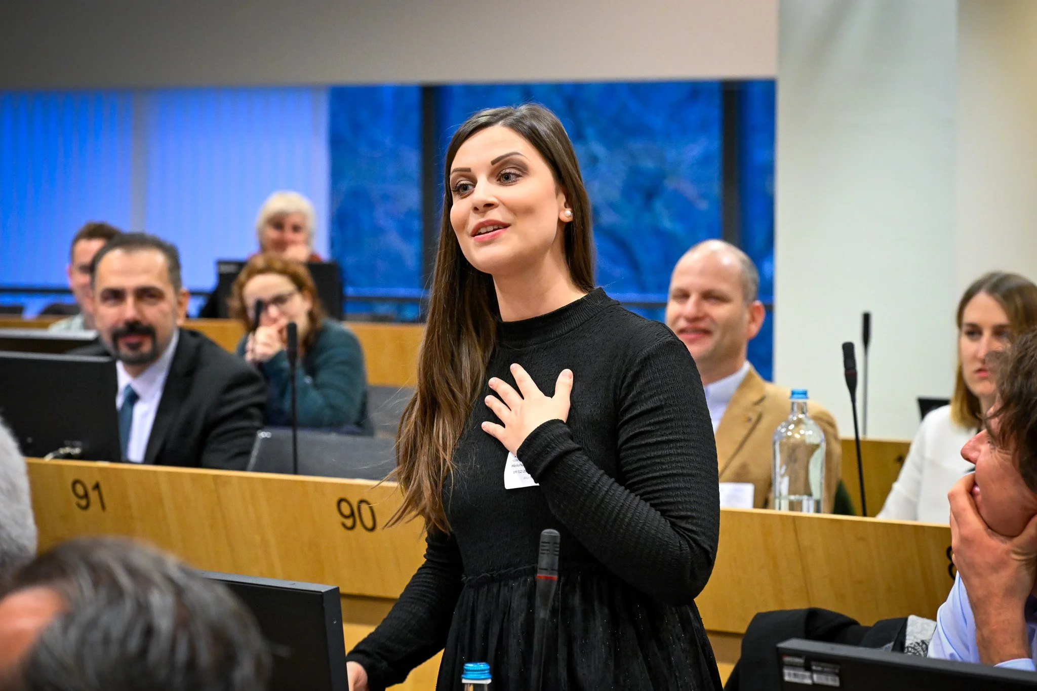 Une femme s'exprime dans une salle de conférence, entourée d'autres participants assis.