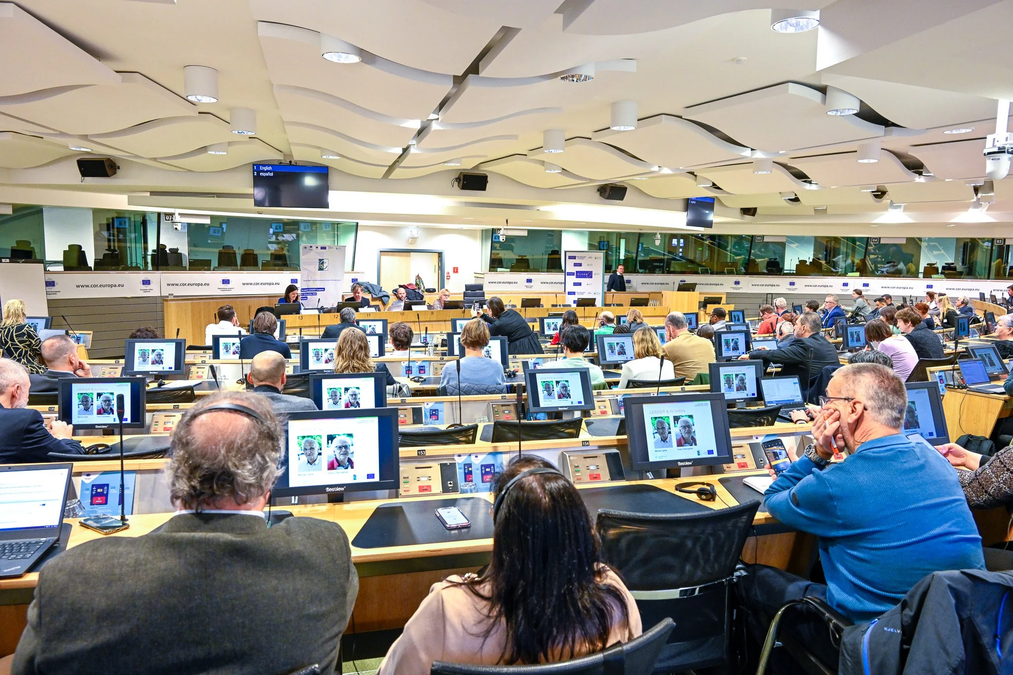Salle de conférence moderne avec plusieurs personnes assises à des bureaux équipés d'ordinateurs, participant à une réunion avec des intervenants à l'avant.