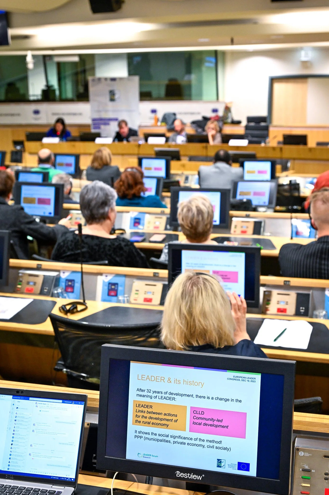 Des personnes assistent à une conférence dans une salle avec des écrans affichant des présentations sur le programme LEADER, axé sur le développement rural et local.