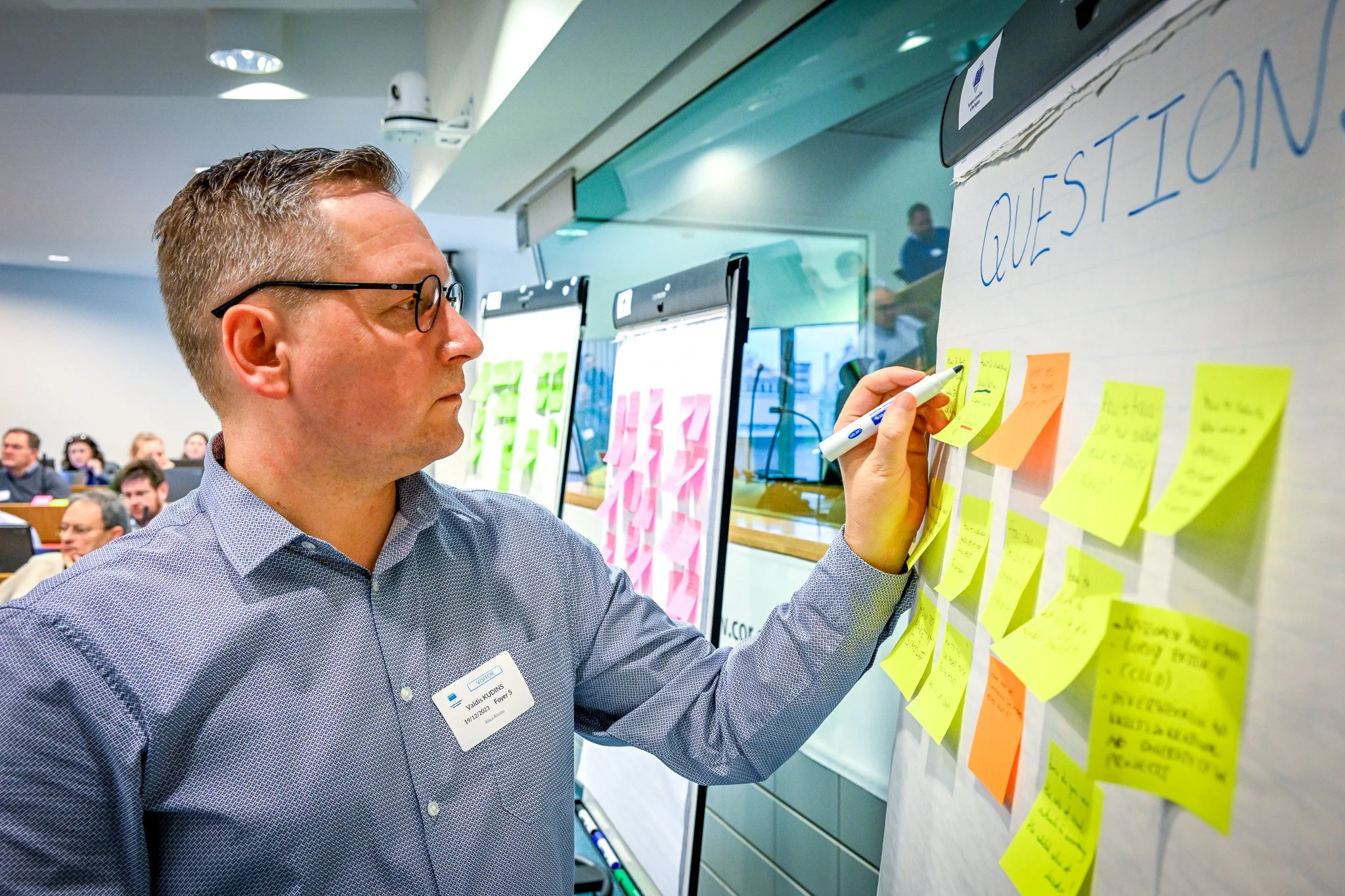 Une personne écrit sur des post-its jaunes attachés à un tableau lors d'une réunion, avec d'autres participants en arrière-plan.
