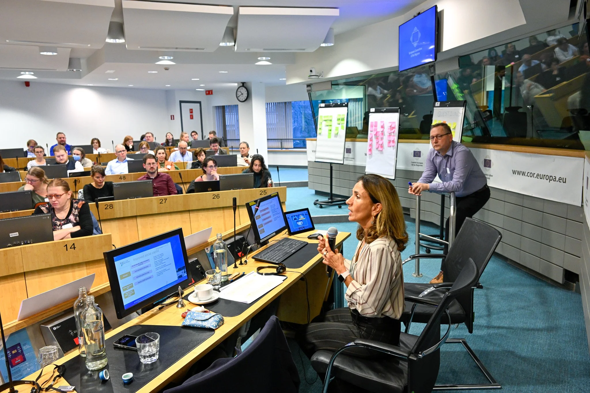 Salle de conférence avec des participants assis, un présentateur tenant un micro, des écrans d'ordinateur, des tableaux avec post-its colorés, et une horloge murale. Environnement professionnel et éducatif.