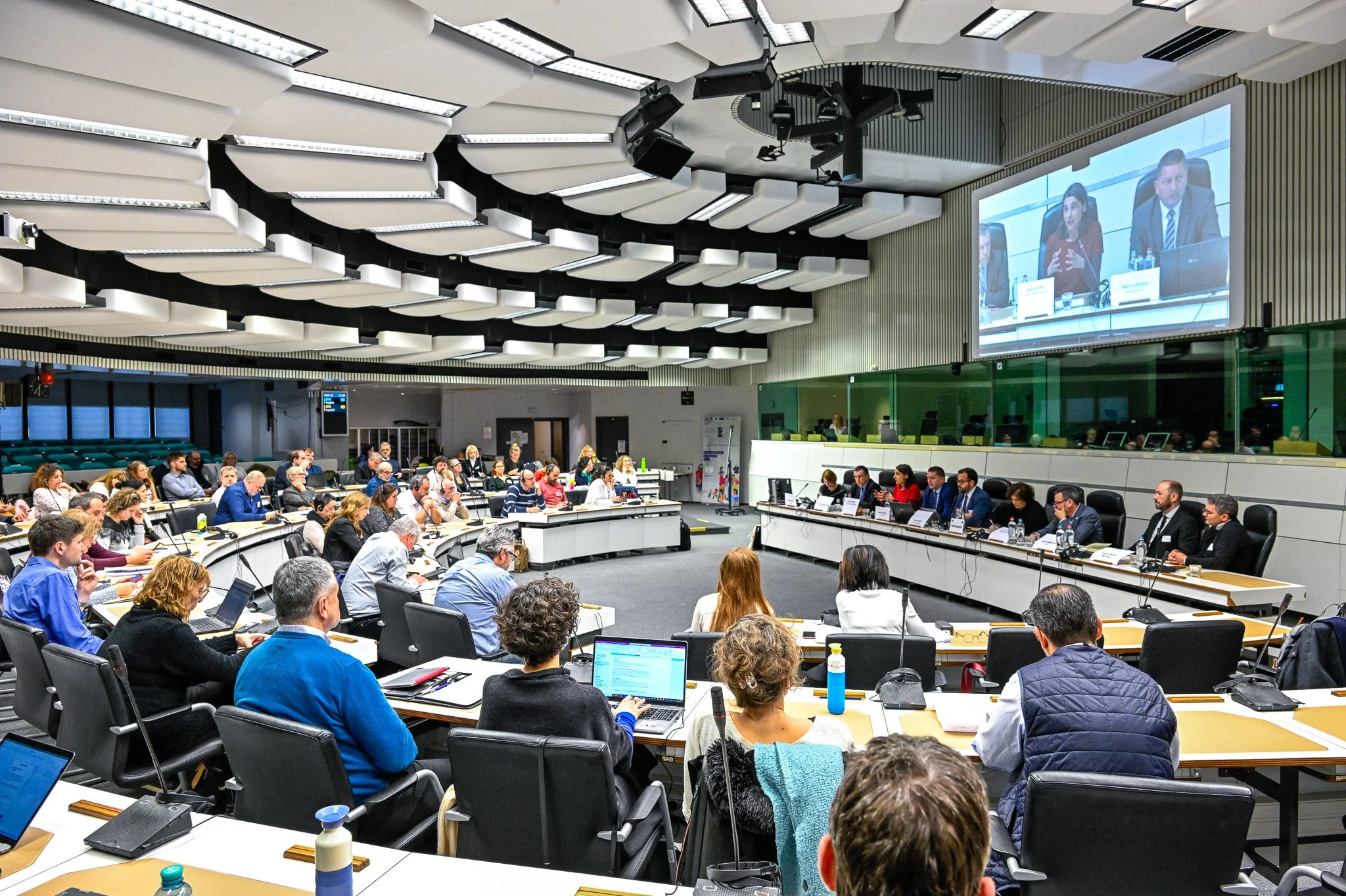 Réunion dans une grande salle de conférence avec des participants assis à des tables, un écran de projection montrant une présentation en arrière-plan.